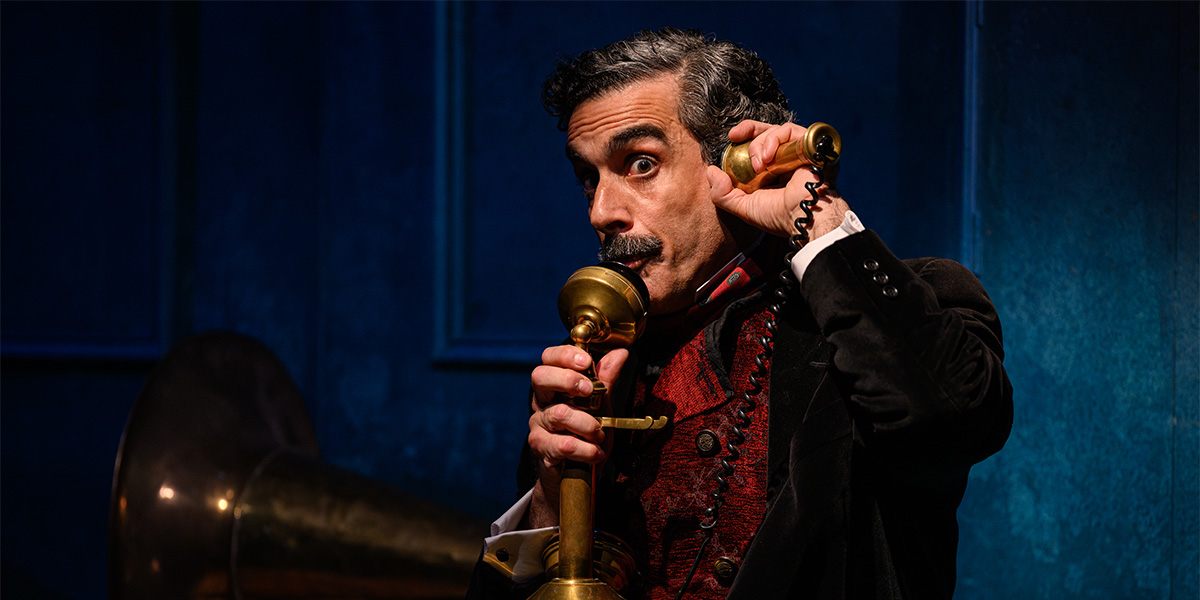 Person in a red vest and dark jacket making a puzzled expression while handling an old-fashioned telephone in a vintage blue room.