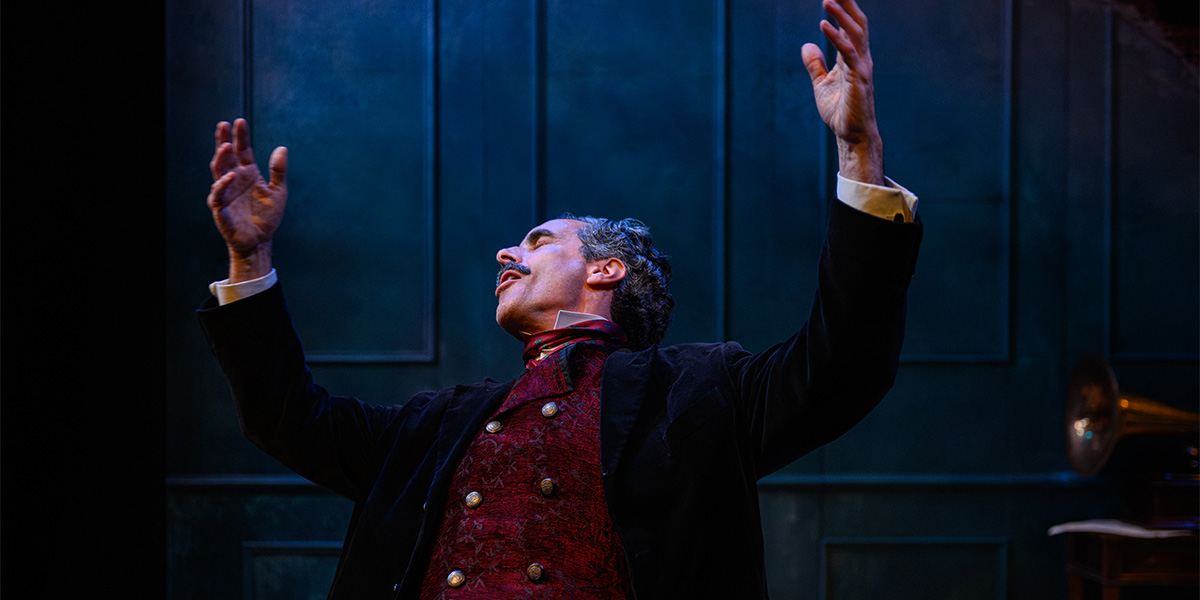 A person performs on stage in a theatrical setting, dressed in a period costume with a dark red vest and black coat, arms raised and expression passionate. The backdrop features a deep blue, panelled wall.