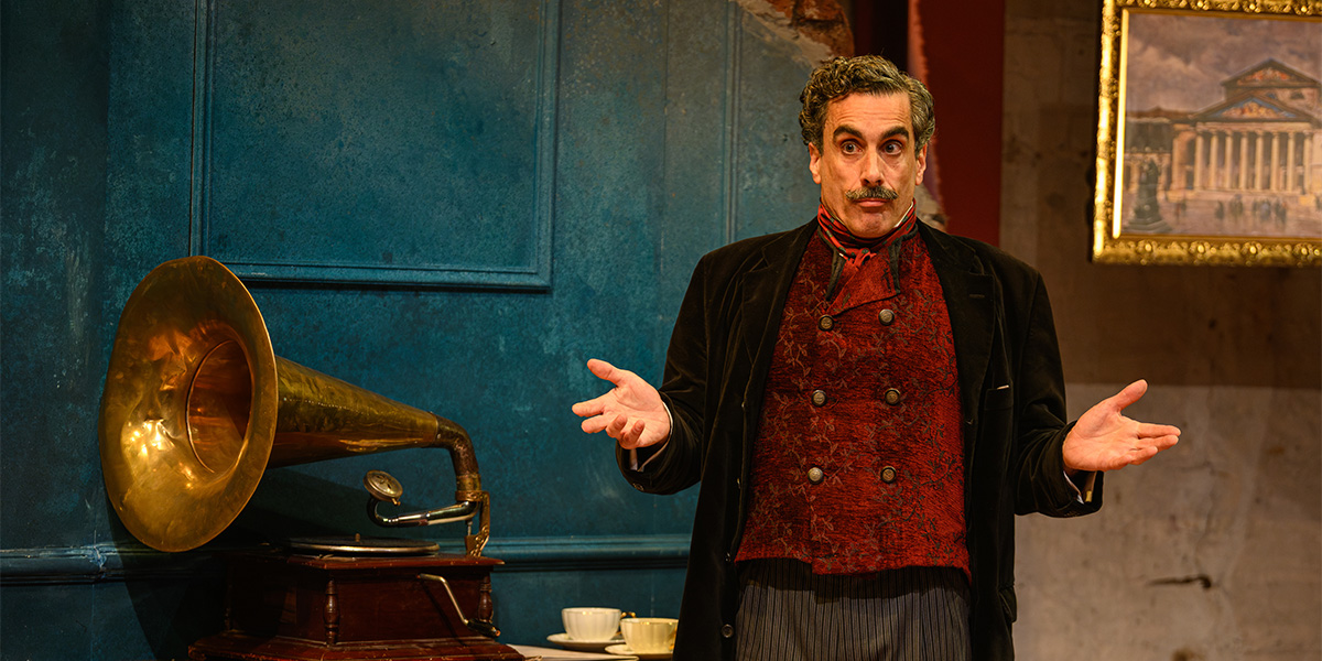 A person stands by a gramophone on a table, gesturing with hands open in a theatrical setting, wearing a formal vest and jacket. The background includes a blue wall and a framed picture.