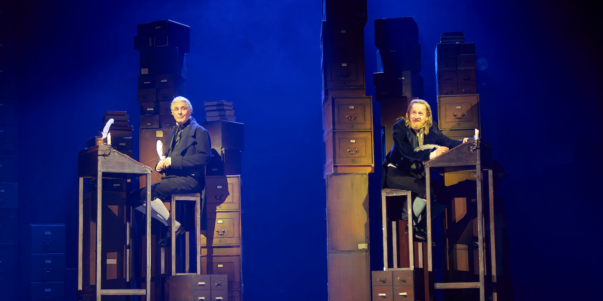 Two actors sit at desks surrounded by tall stacks of wooden drawers, illuminated by blue stage lighting.