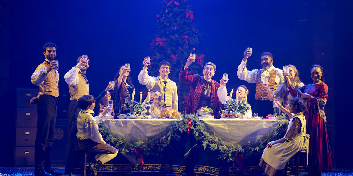 A scene from A Christmas Carol featuring actors in Victorian costumes sitting at a large table. They are raising a toast.