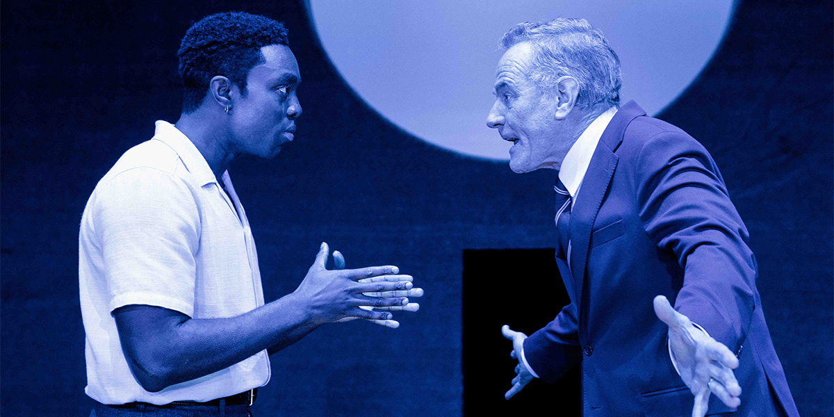 Paapa Essiedu as Chris Keller and Bryan Cranston as Joe Keller, both in formal attire, on stage.