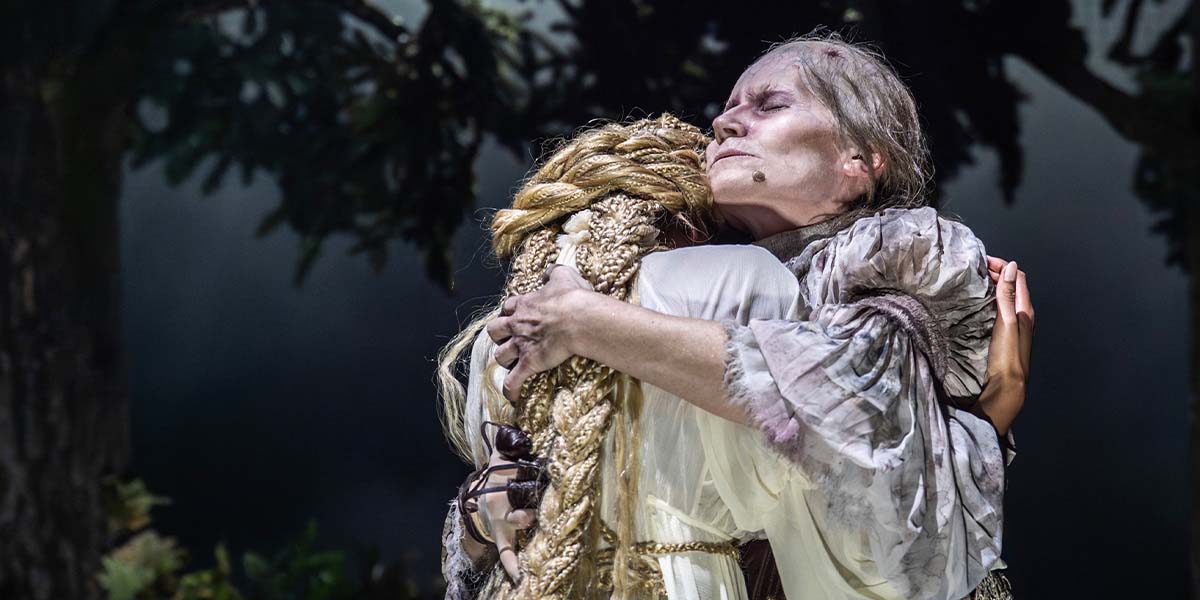 Bella Brown and Kate Fleetwood pose together, smiling, in costume for "Into The Woods," with a forest backdrop.