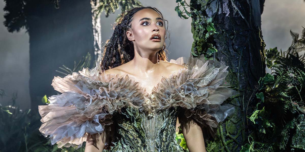 Chumisa Dornford-May stands in a forest, wearing a green dress, surrounded by lush foliage and sunlight.
