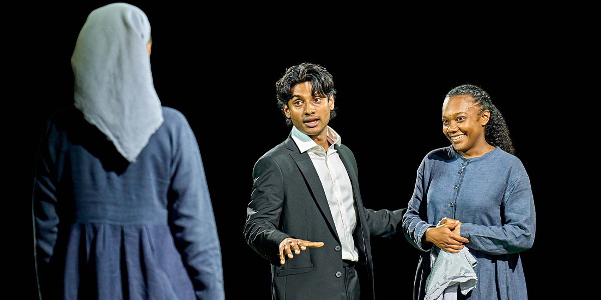 Three actors on stage in a theatrical production, with one wearing a hooded garment, the middle one in a suit and gesturing with an extended hand, and the third smiling and wearing a casual shirt and pants.