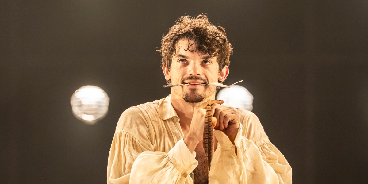 A close-up of a person with short dark hair, smiling, showcasing prominent teeth against a blurred background.