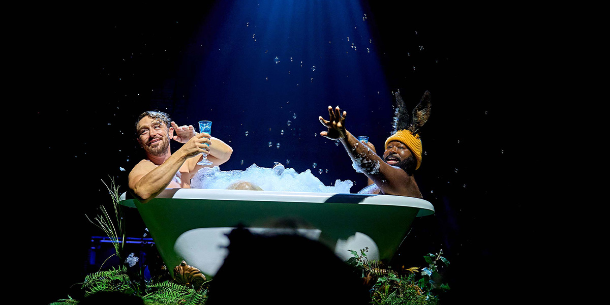 Two individuals enjoying a theatrical bubble bath onstage, one holding a drink, under a spotlight, surrounded by foliage.
