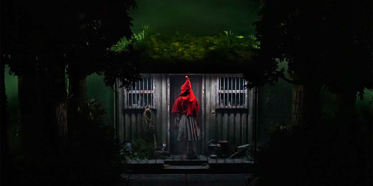 A figure in a red cloak stands before a rustic cabin, surrounded by dark trees and foliage.