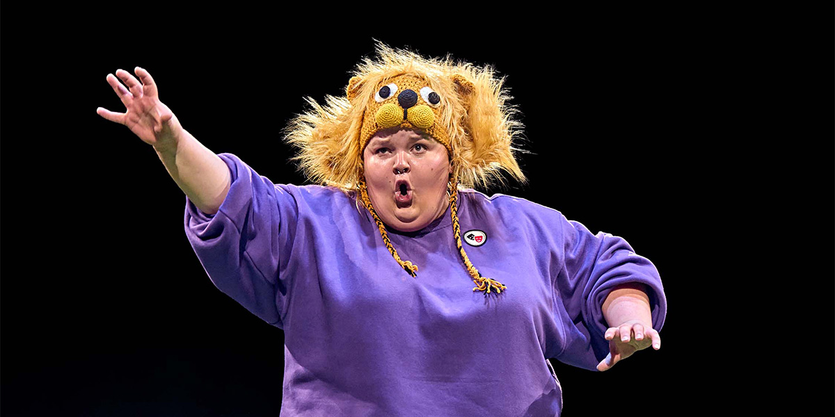 Person performing on stage wearing a lion hat and a purple shirt, gesturing dramatically.