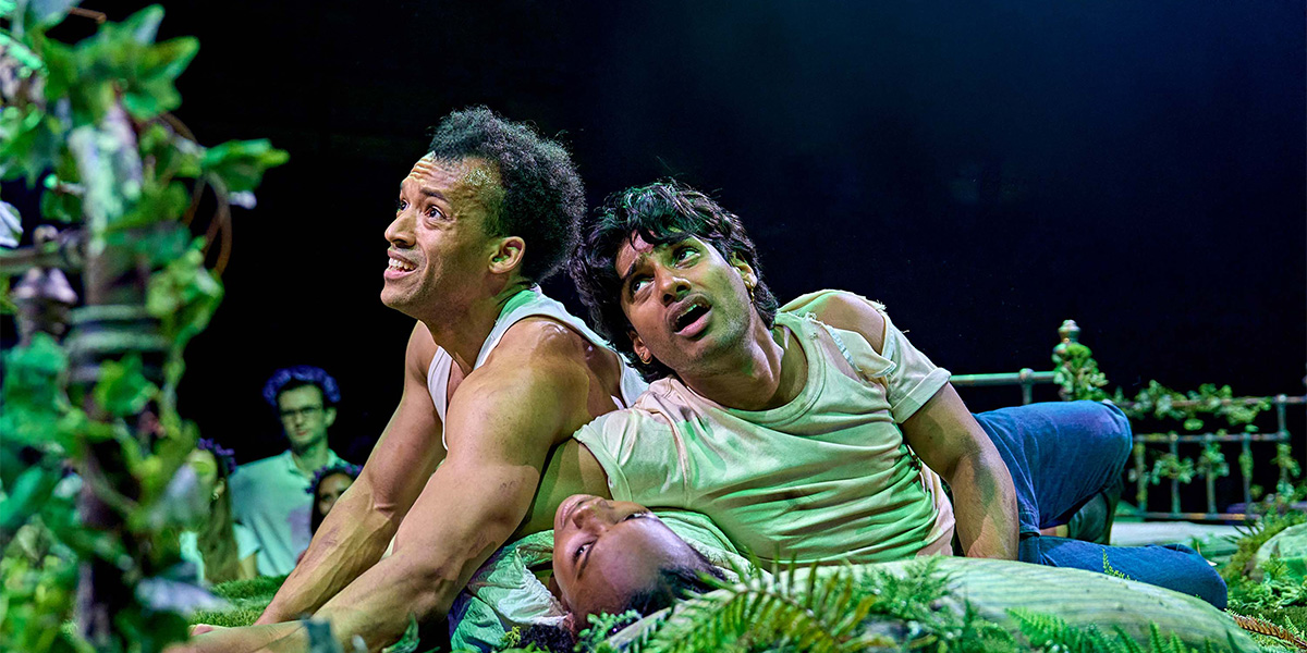 Two actors express shock and concern in a vivid theatrical scene set in a lush, plant-filled environment.