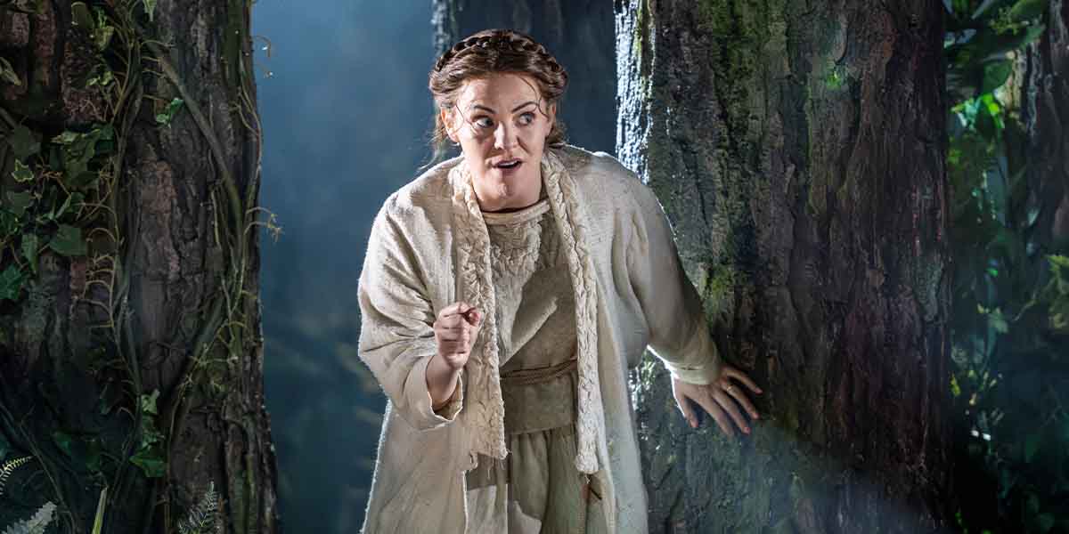 Rachel Tucker (Baker's Wife), dressed in a light-coloured costume, gestures expressively against a forest backdrop.