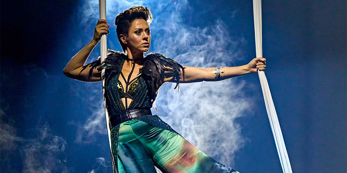 Performer in an elaborate costume performing an aerial silk act against a backdrop of dramatic blue lighting and smoke.