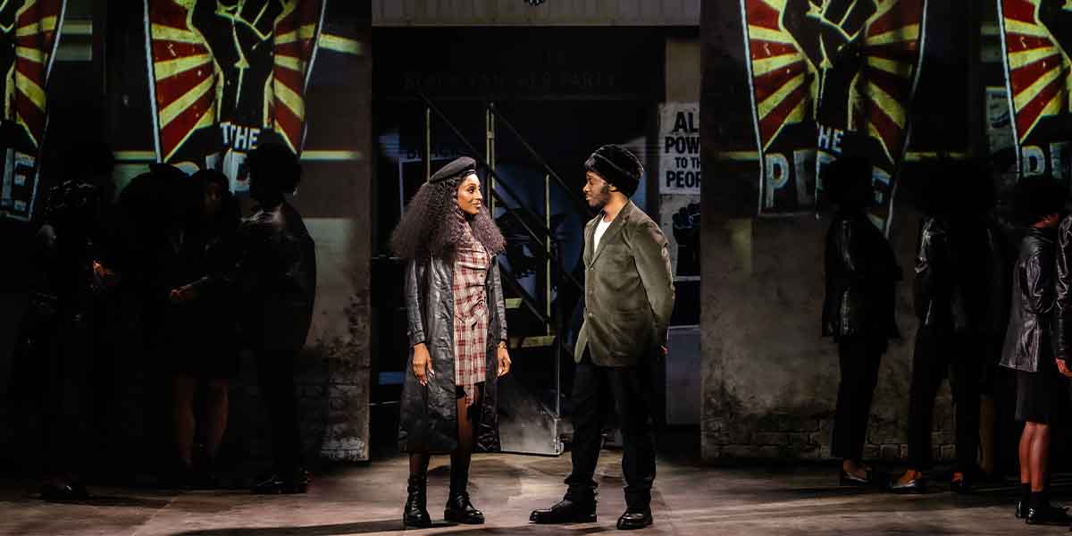 Two performers stand facing each other, surrounded by dramatic posters and a dimly lit background.