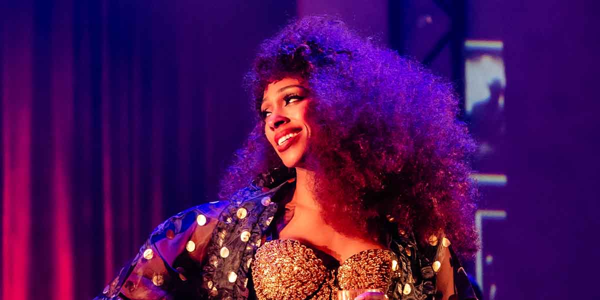 A performer with curly hair smiles, wearing a sparkling gold outfit and a sheer black jacket.