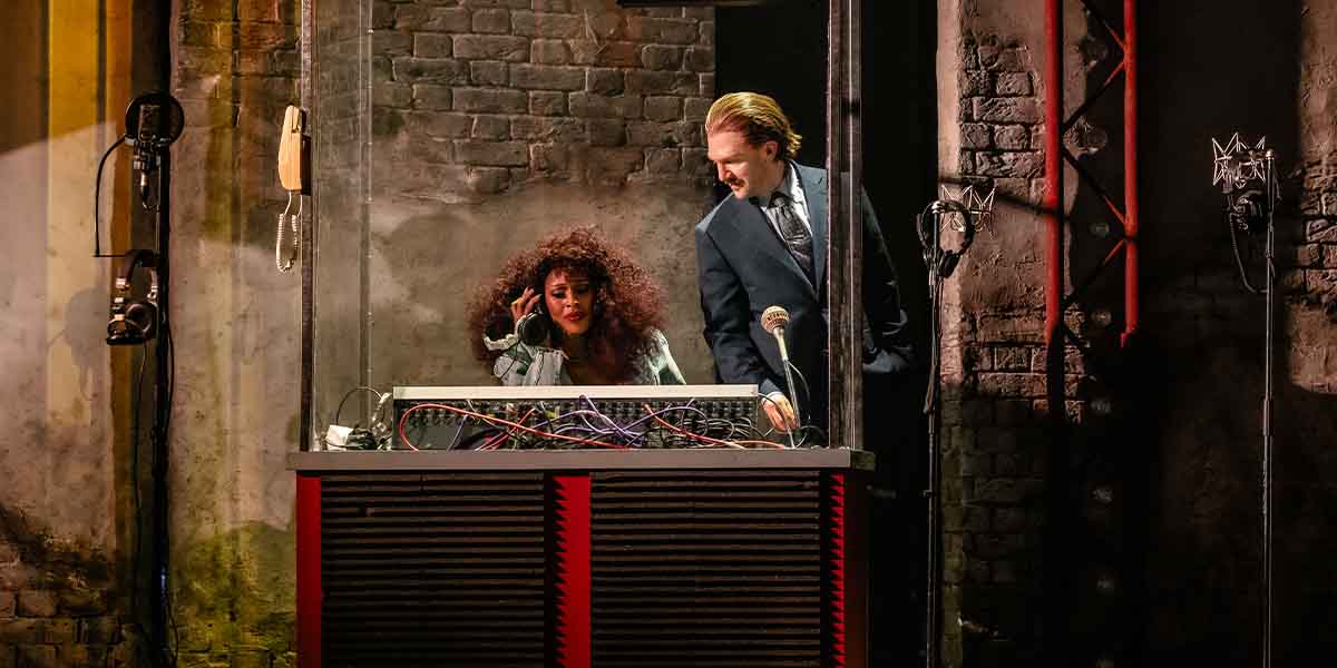 A person with curly hair speaks into a vintage phone while another adjusts equipment in a studio.