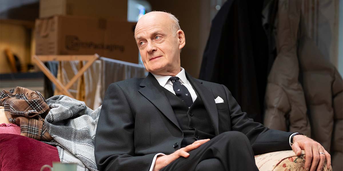 A man in a suit stands with a concerned expression, holding a microphone against a blurred background.