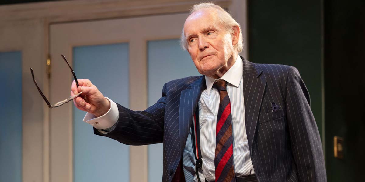 An elderly man in a pinstripe suit holds glasses, looking thoughtfully towards the audience against a green backdrop.