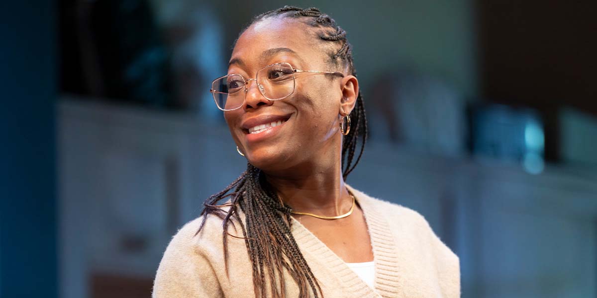 A woman with braided hair and glasses smiles, wearing a beige sweater, against a blurred background.