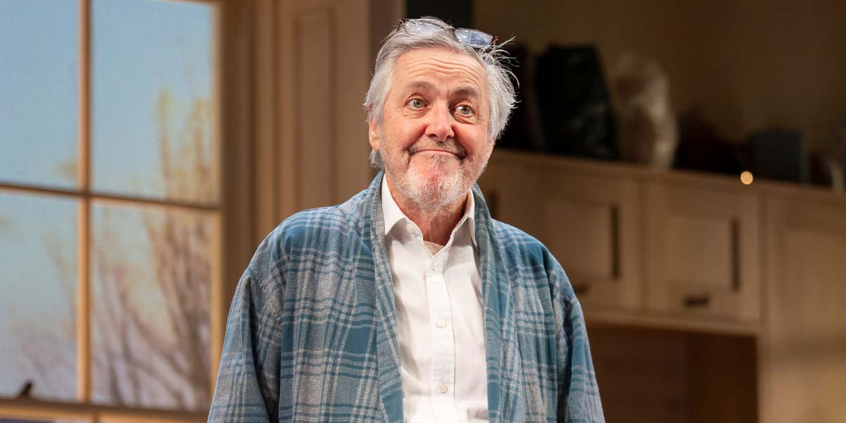 A man with grey hair smiles, wearing a blue plaid robe over a white shirt, indoors.