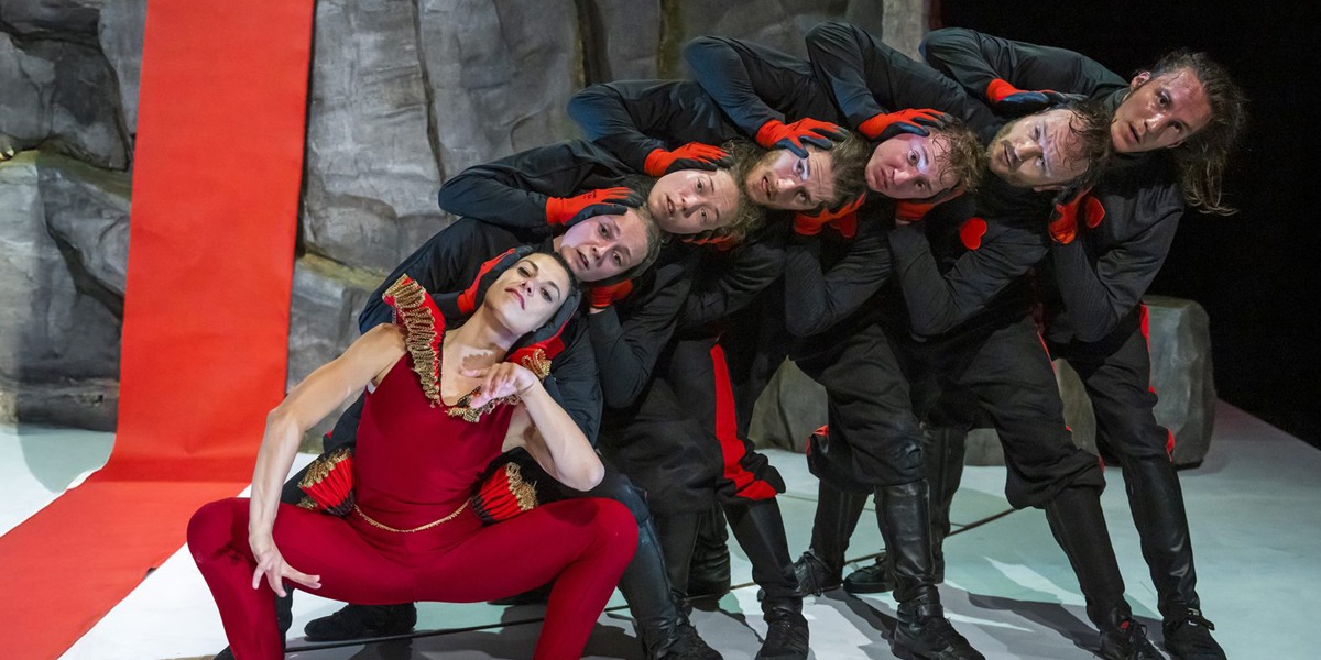 Seven dancers are lined up, from crouched to standing, with their heads stacked and curving in a line. The front dancer wears a red leotard with regal trim, while the others are dressed as soldiers with red gloves. Behind them, a jagged rock wall and red carpet span across the stage.