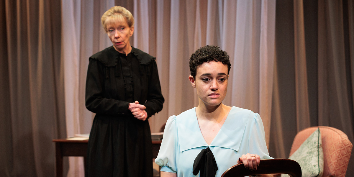 Angela Sims stands in a black dress, while Lily Nichol, in a light blue blouse, looks distressed.