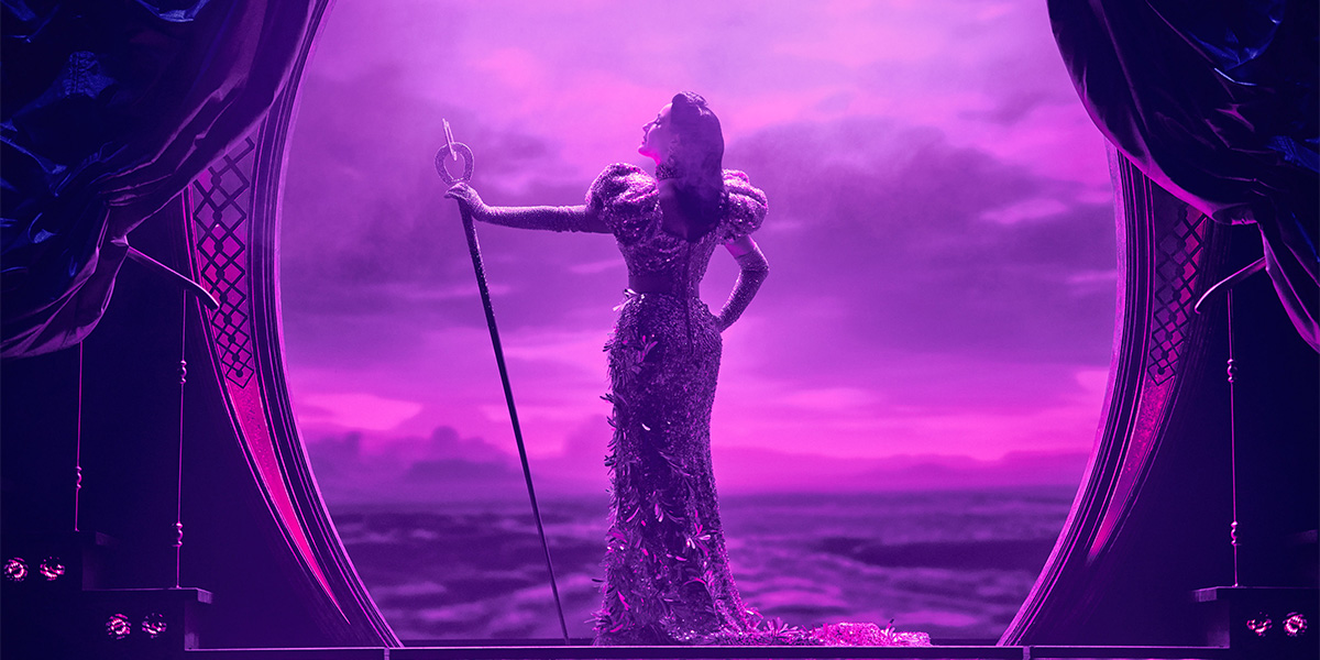 An elaborately dressed character stands on a theater stage set against a dramatic ocean backdrop under a purple sky, holding a scepter.