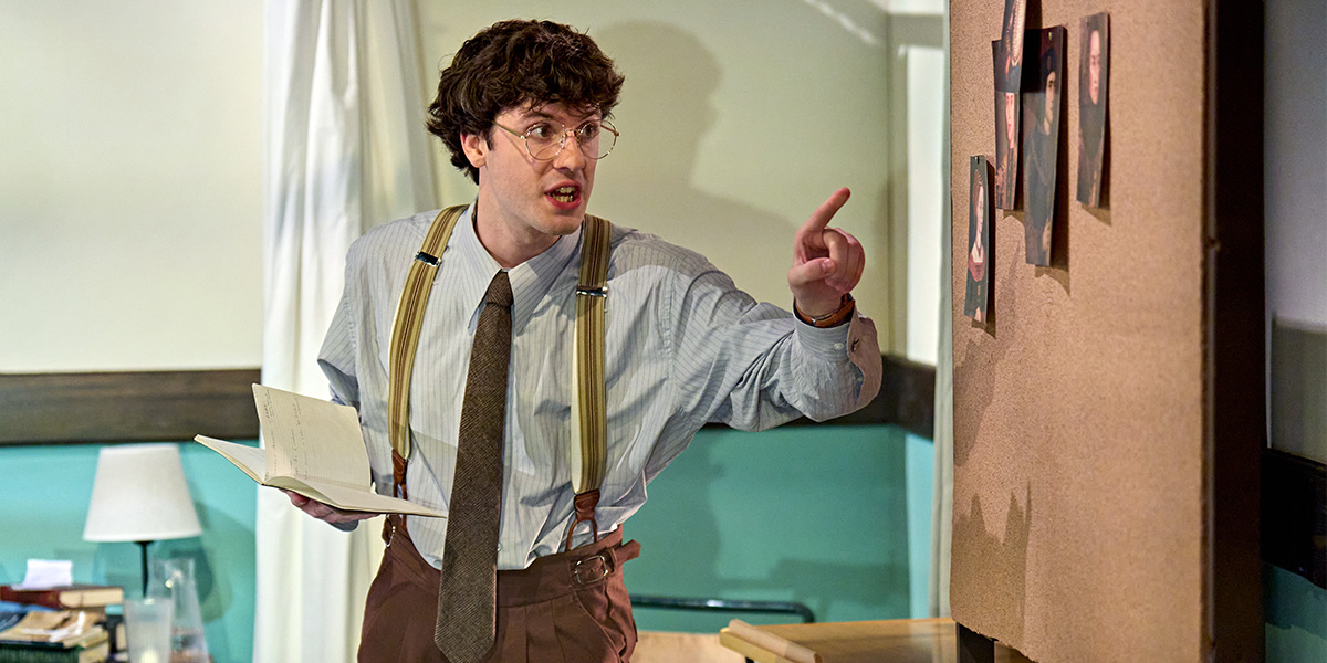 Person with an expressive gesture, holding papers and pointing to a bulletin board, dressed in vintage style with suspenders and glasses.