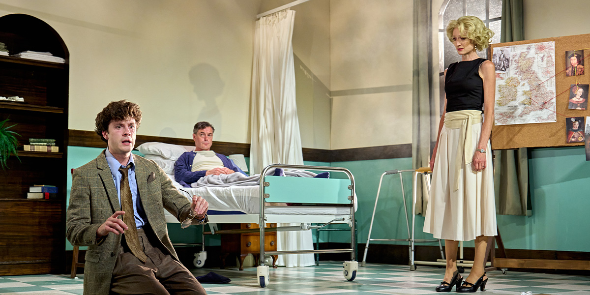 A theatrical scene with three characters on stage; one is seated on a hospital bed, another stands near a bulletin board filled with photos and notes, and the third is seated in the foreground, wearing a suit and looking distressed. The setting suggests a mid-20th century hospital room.