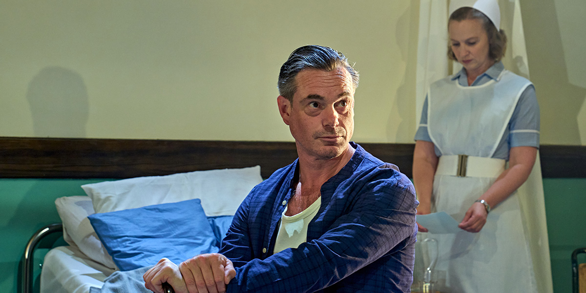 A person sitting on a hospital bed in a blue robe with a concerned expression, while a nurse stands in the background.