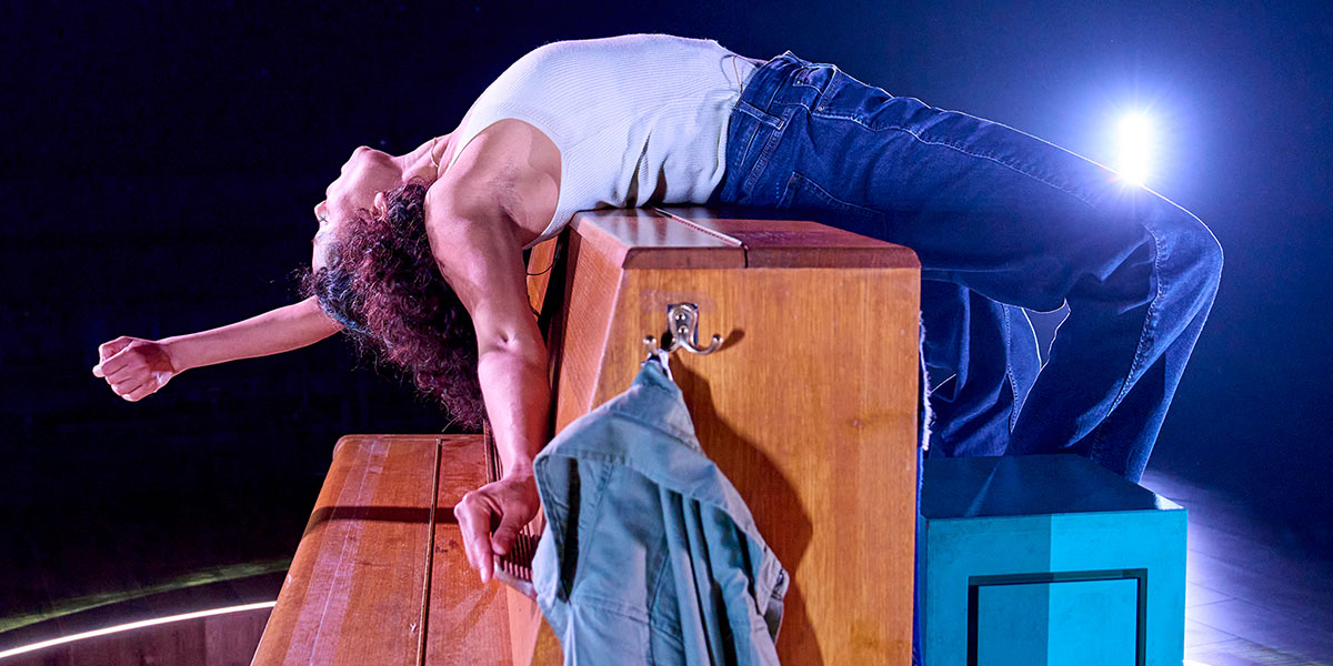 A person performs an expressive dance move, arching backward over a wooden piano on a stage with a spotlight in the background.