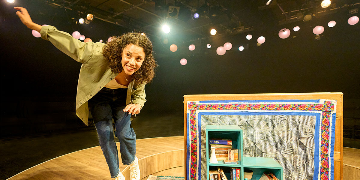Performer smiles while leaning forward on a colourful stage set, under hanging pink lights.