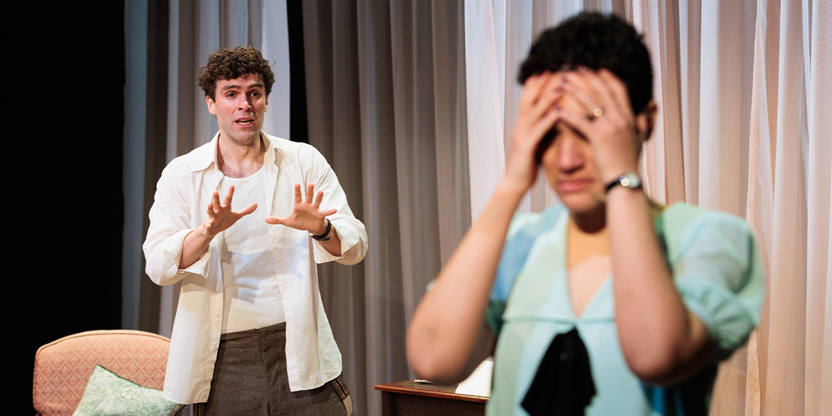 Ewan Miller and Lily Nichol engage in a dramatic scene looking visibly upset, in a vibrant setting with colourful decor.