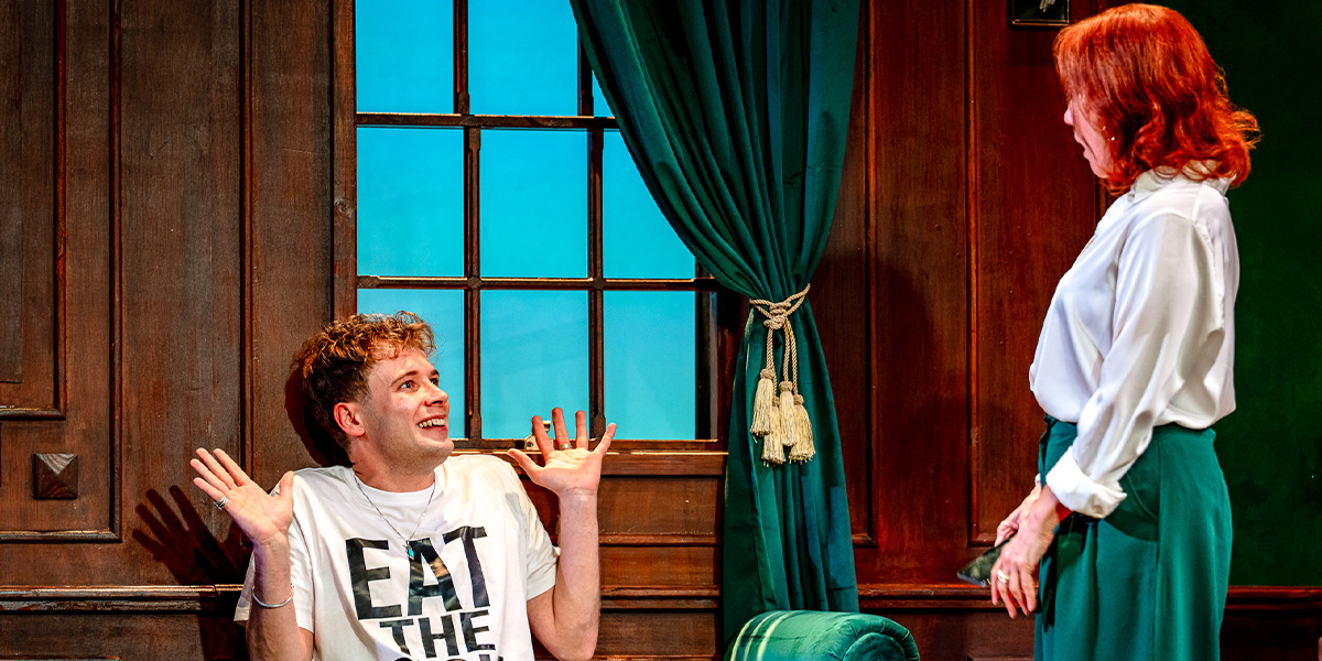 Two actors on a stage set resembling a living room, one seated and the other standing, engaging in a lively conversation. The seated actor wears a T-shirt with the slogan "EAT THE RICH," and the standing actor is dressed in a green pencil skirt and white blouse.