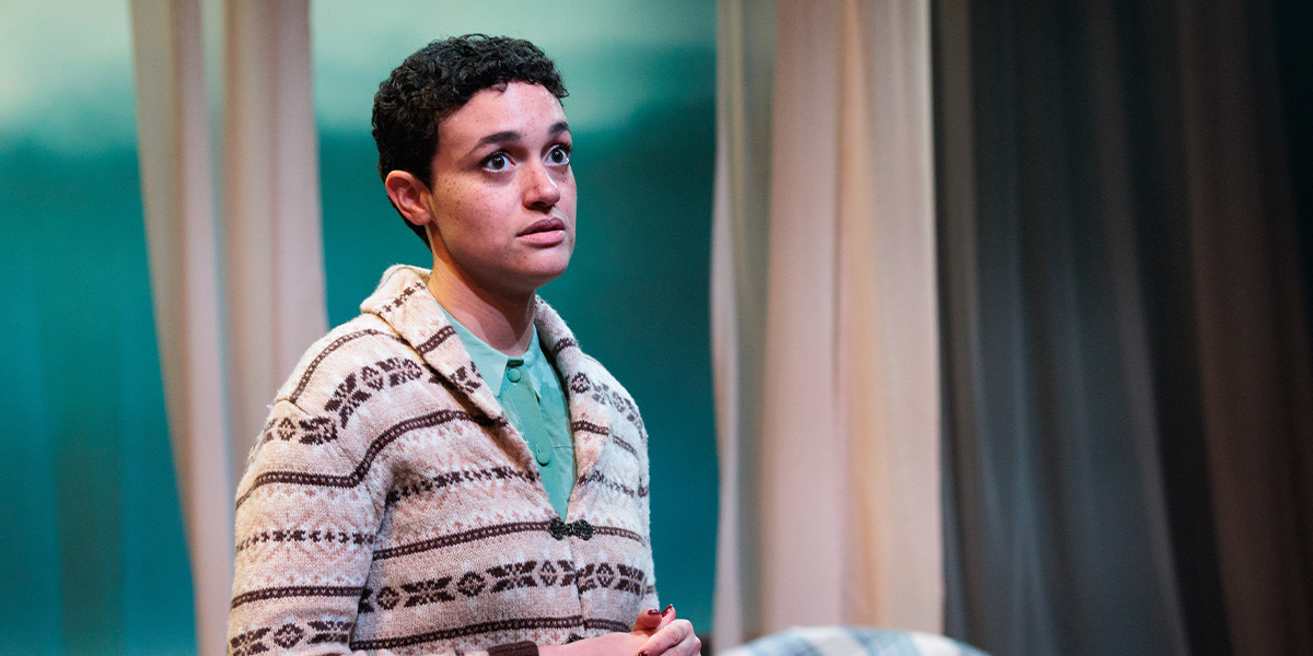 Lily Nichol stands confidently, looking to be deep in thought, with a blurred background featuring soft lighting and neutral tones.