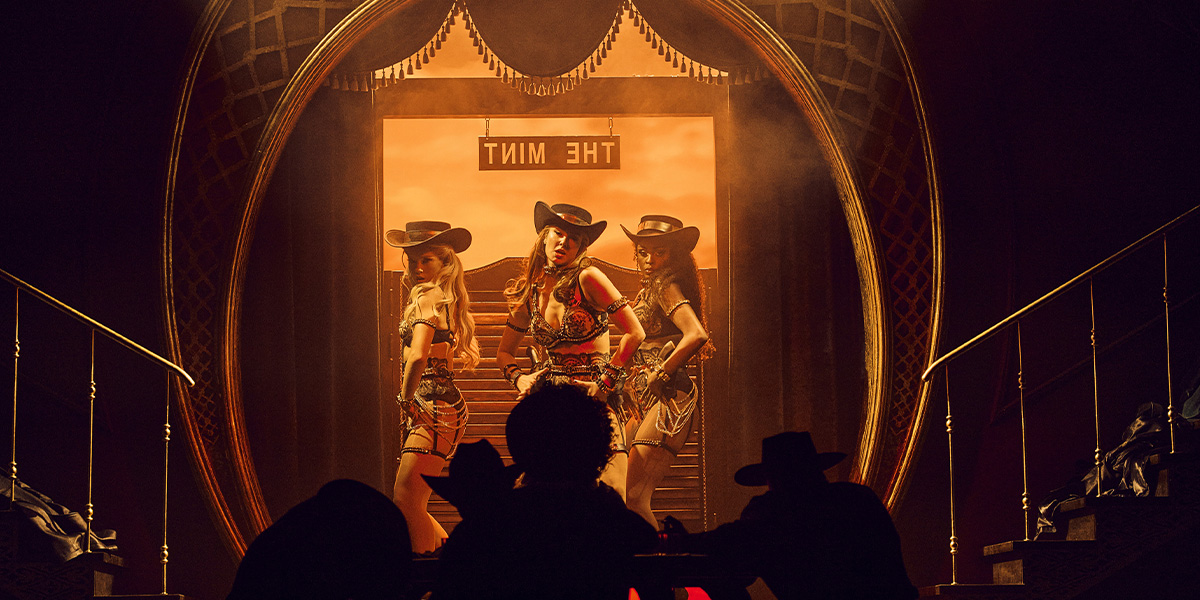Three performers on cowboy-themed stage, framed by dramatic lighting and an ornate backdrop, with silhouettes of audience members in the foreground.