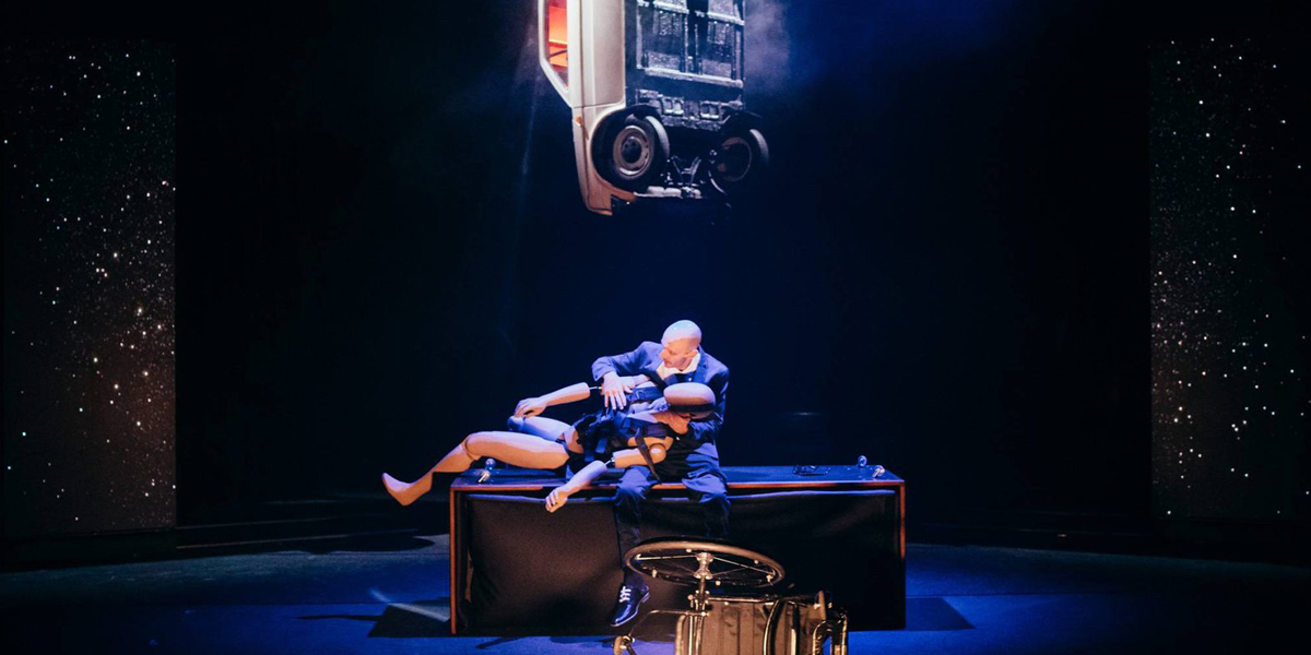 A performer is lying on a table on stage holding a crash test dummy, there is a car suspended above, creating a dramatic illusion, set against a starry backdrop.