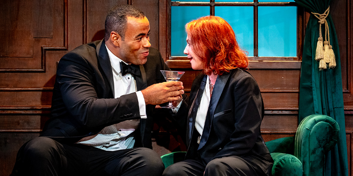 Two individuals in business attire toast with drinks while sitting closely on a couch, set against a backdrop of a room with classic decor.