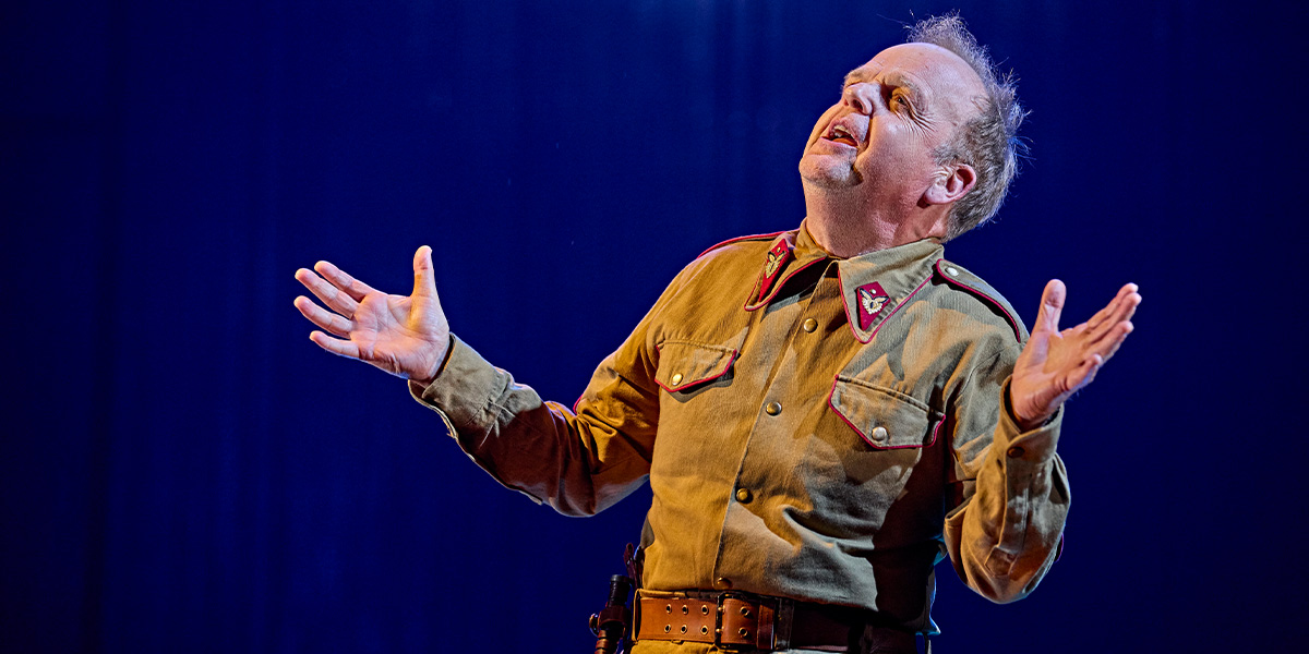 Toby Jones as Iago, wearing dark clothing, with a serious expression, against a dimly lit backdrop.
