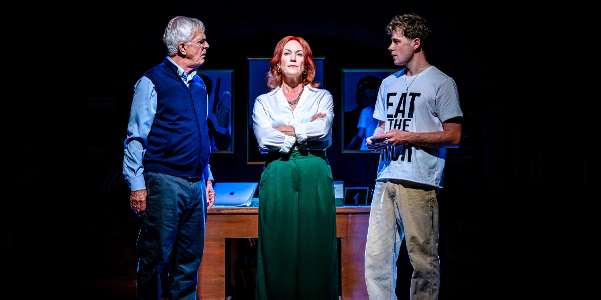 Three actors on stage in a dramatic scene, with one standing behind a desk and the other two facing each other in discussion.