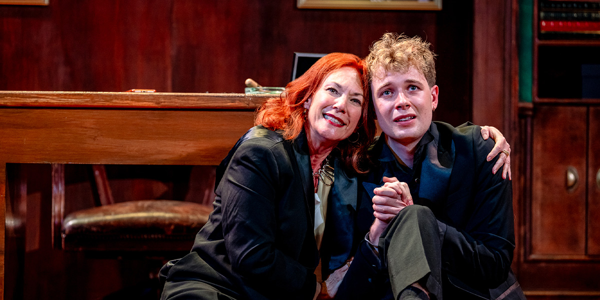 Two actors performing a scene on stage in a theater production, with one person resting their head affectionately on the other person's shoulder, both sharing a moment of connection. The setting includes detailed wooden furniture suggesting a cosy interior space.