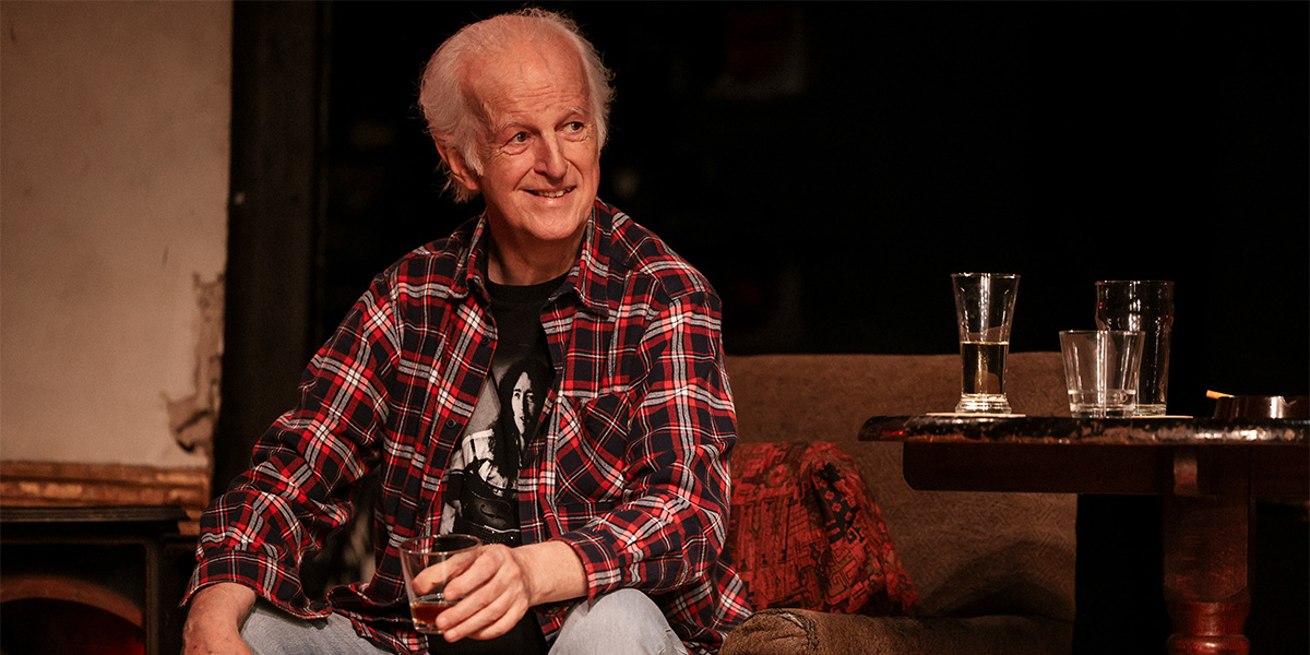 Two men in casual clothing sit on a wooden bench, engaged in conversation, with a rustic interior background.