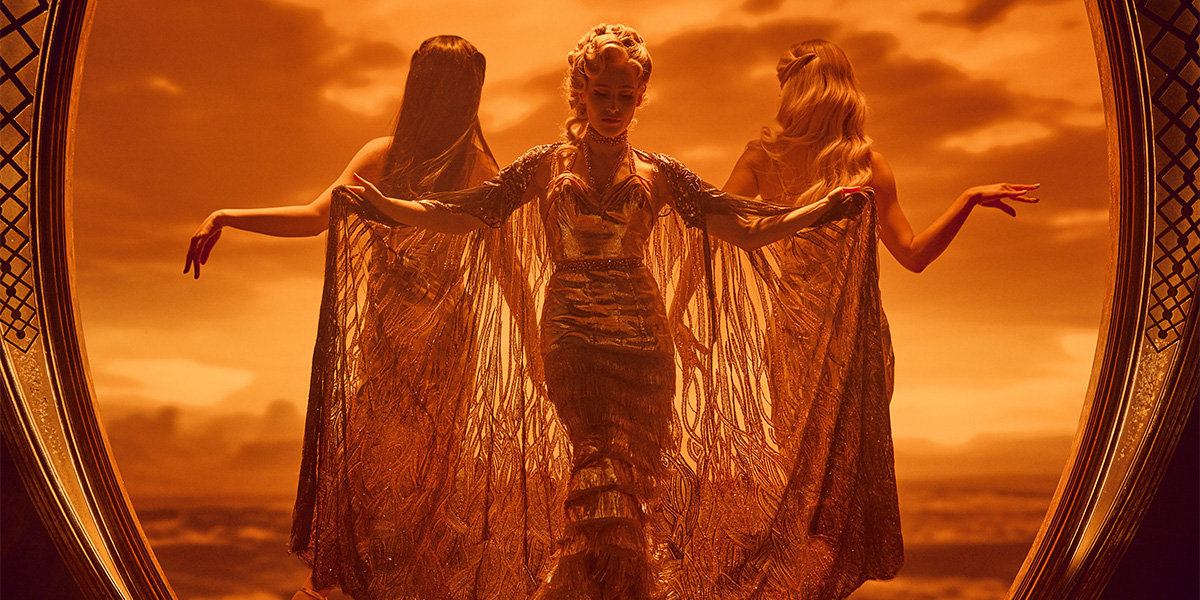 Three characters dressed in elaborate costumes and striking dramatic poses against a vivid, orange-hued desert backdrop.