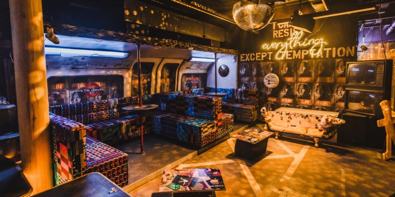 A colourful lounge area with patterned seating and a neon sign in a dimly lit space.