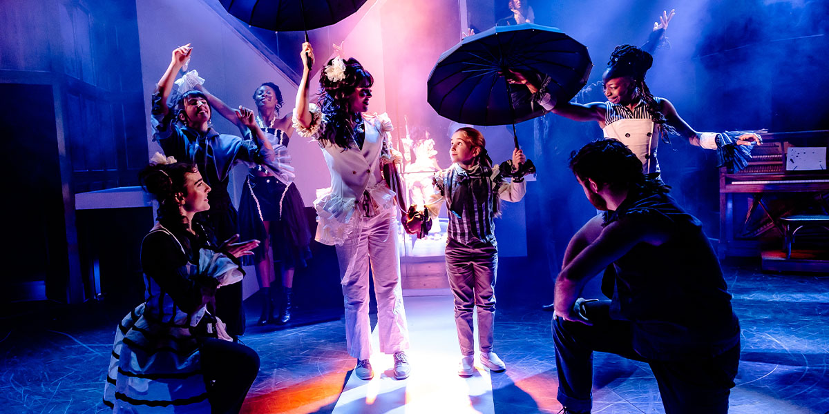 A dynamic theatre scene with multiple performers in colourful costumes engaging in expressive actions on stage, highlighted by dramatic lighting and vibrant set design.
