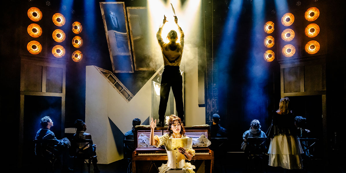 A theatrical performance featuring a central character in a spotlight performing on stage with other actors seated around, set against a backdrop of bright stage lights and a darkened setting.