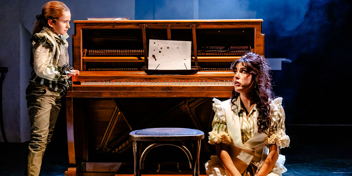 Two actors on stage in a dramatic scene, one standing beside a piano with sheet music on it and the other seated on a stool, both dressed in historical costumes under stage lighting.