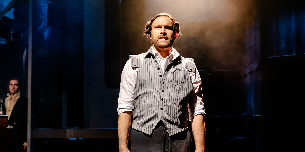 Person on stage performing in a theatrical setting, wearing a vest and striped shirt, with intense lighting highlighting the scene.