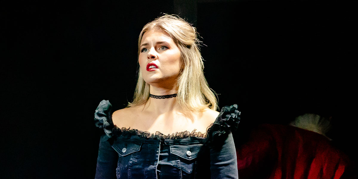 A person performing on stage, dressed in a dark, frilly outfit and a choker, appears under a spotlight with a focused expression.