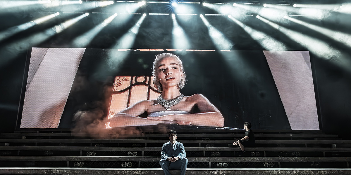 A person sits on stage steps beneath a large screen illuminated by dramatic overhead lighting.