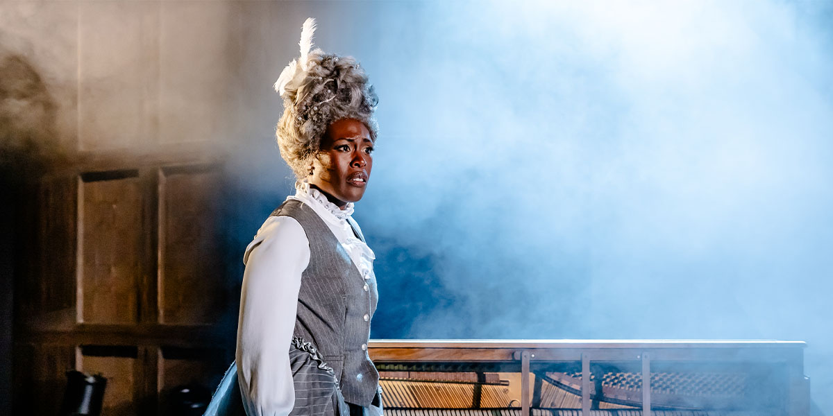 A performer wearing a period costume with a white wig stands on stage in a dramatic theatrical setting.